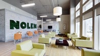 This single-floor space is designed as a contemporary communal lounge or lobby area, blending industrial and modern aesthetics to create a welcoming, functional environment. The most distinctive feature is the eye-catching wall display of large letters spelled out in preserved moss, mounted against an exposed brick backdrop painted in a light grey tone, adding a bold, biophilic focal point to the space.

The ceiling is left exposed, revealing ductwork and structural elements typical of industrial design, while a combination of pendant drum lights and linear fluorescent fixtures ensures the space is well-lit. Large windows along one side of the room invite substantial natural light, enhancing the overall brightness and openness of the interior.

The seating is arranged in multiple groupings, featuring light chartreuse upholstered armchairs and sofas paired with dark wood coffee tables, creating distinct conversation zones. Additional dining-style seating with orange chairs and wooden tables occupies one corner, offering a more casual setting. A wall-mounted flat-screen television is positioned near the center, adding a functional media element. Light hardwood flooring and a natural fiber area rug complete the clean, contemporary aesthetic.