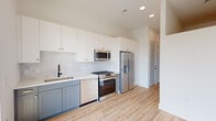 Located at 1015 Boulevard SE in Atlanta's Grant Park area, this single-floor unit showcases a thoughtfully designed, modern interior with consistent light hardwood flooring and a crisp, neutral palette throughout. The open-concept kitchen is a standout feature, outfitted with a two-tone cabinetry scheme—white upper shaker-style cabinets with brass hardware paired with soft gray-blue lower cabinets. White quartz countertops, a white tile backsplash, a matte black gooseneck faucet, and a full suite of stainless steel appliances, including a gas range, built-in microwave, dishwasher, and French door refrigerator, complete the space. Recessed lighting illuminates the kitchen and hallway areas efficiently. The living area opens to a private balcony through floor-to-ceiling sliding glass doors, offering elevated treetop and cityscape views that flood the space with natural light. The bathroom continues the home's modern aesthetic with light wood-look flooring, gray shaker-style vanity cabinetry, white quartz counters, matte black fixtures, and a fully tiled soaking tub and shower surround. Clean white walls and minimalist door hardware with black accents tie the overall design together cohesively.