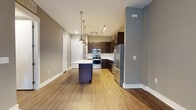 This well-appointed single-floor unit showcases a thoughtfully designed open-concept layout that seamlessly connects the kitchen and living areas. The kitchen is a standout feature, outfitted with rich dark espresso cabinetry, crisp white countertops, and a full suite of stainless steel appliances including a refrigerator, range, microwave, and dishwasher. A subway tile backsplash adds a clean, contemporary touch, while a central island with pendant lighting overhead provides additional prep space and a natural focal point. Warm hardwood-style flooring runs throughout the main living area, lending a cohesive and inviting feel to the space. The bedroom is generously sized with large windows that fill the room with natural light and offer views to the exterior. Dark hardwood-style flooring grounds the room with an elegant contrast to the neutral wall tones. A bathroom connects to a dedicated walk-in closet with built-in shelving and hanging rods, offering practical and organized storage. The unit's neutral color palette of warm taupes and soft grays creates a calm, modern ambiance throughout, making it a versatile and functional living space.