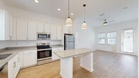 This single-floor space is designed with a clean, modern aesthetic highlighted by crisp white walls, light wood-tone flooring, and an abundance of natural light throughout. The kitchen serves as the centerpiece of the open layout, outfitted with white shaker-style cabinetry, gray countertops, and a full suite of stainless steel appliances including a range, over-the-range microwave, refrigerator, and dishwasher. A freestanding center island provides additional prep and dining space, while two pendant lights overhead add a polished finishing touch. The kitchen flows naturally into an adjoining living area, which features a ceiling fan and windows fitted with horizontal blinds. Additional rooms offer generous wall space, wood-look flooring, and multiple windows that invite natural light. One room includes a glass-paneled exterior door, providing direct outdoor access. The bathroom is well-appointed with white cabinetry, a gray countertop vanity, a large mirror with overhead lighting, and a full soaking tub with a tiled surround. Consistent flooring and a neutral white palette unify the space, creating a cohesive and functional living environment throughout.