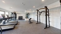 Located at 15 Washington Ave in Carteret, NJ, this modern fitness center offers a thoughtfully designed, single-floor workout space with a generous open layout. The room features neutral-toned walls, recessed lighting, and durable rubber-backed carpet flooring suited for high-traffic exercise use.

Large floor-to-ceiling windows line multiple walls, flooding the interior with abundant natural light while offering views of the surrounding outdoor courtyard and neighboring buildings. The bright, airy atmosphere creates an inviting environment for exercise.

The space is comprehensively outfitted for a full range of workouts. Cardio equipment includes multiple treadmills, elliptical machines, and a stationary cycling bike, all positioned along the windowed walls to take advantage of the outdoor views. A dedicated free weights section features a multi-tiered dumbbell rack with a wide range of weights, stability balls, and kettlebells. Strength training is further supported by a cable machine and a multi-function power rack with an adjustable bench. A large wall-mounted mirror in the strength area enhances spatial perception and allows users to monitor their form. The fitness center is accessible via glass double doors and is designed to support a variety of fitness routines in a clean, modern setting.