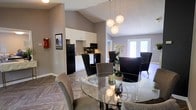 This single-floor space offers an open-concept layout that seamlessly connects the dining area and kitchen, creating a welcoming and functional environment. The dining area is a standout feature, centered around a round glass-top table surrounded by a mix of sleek black and taupe upholstered chairs, all illuminated by a striking cluster pendant light fixture with globe-shaped shades. The vaulted ceiling adds a sense of height and openness to the space, while light wood-look flooring in a herringbone pattern anchors the room with warmth and texture. The kitchen is efficiently designed with white cabinetry, black appliances including a full-size refrigerator, microwave, and range, and clean countertops that offer ample workspace. A set of French doors at the far end of the space floods the interior with natural light and hints at outdoor access, enhancing the overall brightness and airy feel. Neutral gray walls throughout provide a cohesive and modern backdrop, complemented by thoughtful decorative touches such as fresh floral arrangements. The layout and design make this space well-suited for both everyday use and hosting, combining practicality with a refined aesthetic.