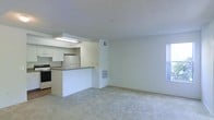 This single-floor space offers a clean, functional layout filled with natural light. The open-concept design connects the kitchen and living area, creating a cohesive and airy environment throughout.

The kitchen is thoughtfully appointed with white cabinetry, a stainless steel refrigerator, an electric range, and a built-in overhead microwave or range hood. A peninsula with a laminate countertop serves as a natural divider between the kitchen and the living area, providing additional workspace and casual seating opportunities.

The living area features neutral wall-to-wall carpeting that adds warmth underfoot, while bright white walls and a textured ceiling contribute to the space's clean, understated aesthetic. A large window on the far wall floods the room with natural light, enhancing the sense of openness. Visible HVAC vents are integrated into the walls, ensuring efficient climate control throughout.

Overall, the space presents a practical and welcoming environment, characterized by its light color palette, open layout, and well-organized kitchen design — well-suited for comfortable everyday living.