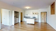 This single-floor space features an open-concept layout that combines the kitchen and living areas seamlessly. The kitchen is outfitted with white cabinetry, dark countertops, a stainless steel sink, a microwave, and a full-size refrigerator. A freestanding island with a dark countertop provides additional workspace and storage, positioned centrally to maximize functionality. Light hardwood flooring runs throughout the space, contributing to a bright and clean aesthetic. The walls are finished in neutral white, complementing the natural light that fills the room. A dedicated laundry area is tucked into an open closet along one wall, housing a side-by-side washer and dryer for added convenience. Open shelving is mounted near the kitchen, offering accessible storage or display space. A wood-toned entry door is visible on one side of the space, while additional doorways lead to other areas of the unit. The flush-mount ceiling light provides functional overhead illumination. Overall, the space presents a practical, minimalist design with a cohesive color palette of white, light wood, and dark accents.