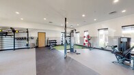 This modern fitness center is thoughtfully designed to accommodate a full range of workout needs across a single, open floor plan. The space is divided into distinct functional zones, each tailored to a specific type of training. The cardio area features multiple treadmills, a stationary bike, and an elliptical machine, all positioned along a wall of large windows that flood the room with natural light and offer views of the surrounding desert landscape with mature cacti and trees. Adjacent to the cardio zone is a dedicated strength and free weights area, complete with a multi-station cable machine, a full dumbbell rack ranging across multiple weight increments, an adjustable weight bench, and a barbell rack set atop green turf flooring — ideal for functional lifting. A training zone along one wall provides TRX suspension straps, wall-mounted storage for additional equipment, and open floor space for dynamic exercises. A leg press and seated machine round out the strength equipment offerings. The interior features a two-tone wall treatment in neutral white and gray, recessed lighting, durable mixed flooring including carpet, rubber, and turf, and a wall-mounted TV for entertainment. A water fountain and restroom access are conveniently located within the facility.