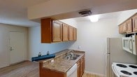 This single-floor space is designed with practicality and an open feel in mind. The kitchen serves as a central feature, showcasing warm medium-toned wood cabinetry with raised panel doors providing ample upper storage. The countertops feature a granite-style surface in earthy tones, complementing the cabinetry. A double stainless steel sink is positioned along the counter, accompanied by a white dishwasher and a white electric range. The kitchen is laid out with a peninsula-style counter that opens toward the adjacent living area, creating a connected and sociable flow between spaces.

The living area, visible from the kitchen, features hardwood-style flooring that extends throughout, adding warmth and continuity to the space. A ceiling fan with a light fixture is installed overhead, providing both air circulation and ambient lighting. The walls feature a textured finish with an accent wall painted in a muted teal tone, offering a touch of color contrast against the neutral surroundings. Overhead vents are integrated into the ceiling, ensuring efficient climate control throughout the unit. Natural light is supplemented by flush-mount and ceiling fixtures.