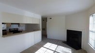 This single-floor space presents a straightforward, functional layout with a clean, neutral aesthetic throughout. The main living area is carpeted and finished in crisp white walls, creating a bright and open atmosphere. Natural light enters through a window, casting geometric patterns across the floor and adding warmth to the otherwise minimal interior. A fireplace with a dark surround serves as a focal point on one wall, offering both visual interest and practical utility.

Adjacent to the living area, a pass-through kitchen counter connects the cooking space to the main room, promoting an open flow between the two areas. The kitchen features light-toned upper cabinetry that extends to the ceiling, maximizing vertical storage. The countertop surface appears to be a neutral laminate, consistent with the space's understated design. A glimpse through a hallway suggests additional rooms beyond the main living space.

Overhead, HVAC vents are integrated into the white ceiling, ensuring climate control throughout. The overall design is minimal and move-in ready, suited for those seeking a clean, uncluttered living environment.