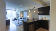 This contemporary single-floor residence offers a thoughtfully designed open-concept layout that seamlessly connects the kitchen and living areas. The kitchen is a standout feature, showcasing sleek dark cabinetry, dark granite countertops, and a central island equipped with a stainless steel sink and integrated dishwasher. Elegant pendant lights hang above the island, adding a refined focal point to the space. A white subway tile backsplash provides a clean contrast to the rich, dark tones of the cabinetry. The kitchen flows naturally into the living area, which is finished with warm hardwood flooring and neutral-toned walls that create an inviting, modern ambiance. Recessed lighting complements the overall illumination throughout the space. A large floor-to-ceiling window in the living area floods the interior with abundant natural light and hints at views of the surrounding cityscape. The open layout and cohesive design language throughout the space make it both functional and visually appealing, ideal for everyday living and entertaining alike.