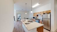 This single-floor residence is designed with a clean, contemporary aesthetic and a practical, open layout. The kitchen serves as a central hub of the living space, featuring warm honey-toned cabinetry, light quartz countertops, stainless steel appliances including a gas range and refrigerator, and a spacious island with an integrated sink that doubles as a breakfast bar. Pendant lighting above the island adds a modern touch, while light hardwood-style flooring extends throughout the main living areas. A dedicated dining area sits adjacent to the kitchen, ideal for casual meals. The living room is bright and inviting, showcasing large windows that frame outdoor greenery and fill the space with natural light. Both bedrooms are finished with plush wall-to-wall carpeting and feature generous closet space. The primary bedroom benefits from direct access to a private en-suite bathroom, which is appointed with white cabinetry, a wide vanity, light stone-toned countertops, and a large framed mirror. A second full bathroom mirrors this clean, neutral design with tile flooring and a white vanity. Throughout the residence, the neutral color palette and consistent finishes create a cohesive and calming ambiance.