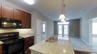 This single-floor space showcases an open-concept layout that connects the kitchen and living area in a cohesive, functional design. The kitchen is a standout feature, centered around a generously sized island topped with light-toned granite countertops that offer both prep space and casual seating potential. A double stainless steel sink is integrated into the island, complemented by elegant pendant lighting suspended above — a trio of bell-shaped fixtures that add a refined decorative touch.

Rich, dark-stained wood cabinetry lines the kitchen walls, providing ample storage while offering a warm contrast to the light granite surfaces. Black appliances, including a range and over-the-range microwave, complete the kitchen's cohesive aesthetic. Under-cabinet lighting enhances the workspace, adding both functionality and ambiance.

The space opens fluidly into the living area, which benefits from natural light streaming in through a set of French doors at the far end. A ceiling fan is positioned centrally overhead, promoting air circulation throughout the open space. Neutral gray walls and light-toned flooring in the living area create a clean, versatile backdrop, while a hallway visible from the kitchen suggests additional private rooms beyond.