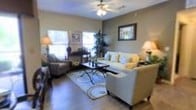 This single-floor residence offers a comfortable and well-designed living environment centered around a spacious main living area. The living room is a standout feature, showcasing rich wood-style hardwood flooring that adds warmth and character throughout the space. Neutral-toned walls create a versatile backdrop, complemented by tasteful wall art and decorative accents that enhance the overall aesthetic.

The living area is generously sized, accommodating a plush sectional sofa and additional seating arranged around a classic wrought-iron coffee table, all anchored by a patterned area rug. A ceiling fan with an integrated light fixture is centrally positioned, providing both functional airflow and ambient lighting. Multiple windows along the walls allow natural light to fill the room, creating a bright and airy atmosphere while offering views of the surrounding greenery.

Lush indoor plants add a refreshing natural element to the space, contributing to a relaxed and homey ambiance. The thoughtful furniture arrangement promotes an open flow throughout the living area, making the space feel welcoming and practical. Overall, this residence combines functional design with warm, inviting details that create a comfortable everyday living experience.