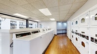 Located at 4333 Chesford Rd in Columbus, Ohio, this single-floor communal laundry facility offers a well-organized and functional space for residents. The room features warm hardwood flooring throughout, complemented by a drop tile ceiling with recessed panel lighting that ensures bright, even illumination across the entire area.

A row of commercial-grade top-load washing machines lines one side of the space, while the opposite side houses multiple stacked dryer units, maximizing the available floor area and providing ample laundry capacity. At the far end of the room, a recessed seating area with a built-in bench provides a convenient waiting space for users.

One of the standout features of this facility is the expansive floor-to-ceiling glass wall along the exterior-facing side. The large black-framed windows and glass panels flood the interior with natural light and offer clear views of the surrounding property grounds, including the outdoor walkways, stairwells, and the neighboring residential buildings. This transparency gives the space an open, airy feel that is uncommon in utility-focused facilities.

The layout is practical and efficient, with a wide central aisle providing easy access to all machines. Overall, the space combines functionality with a clean, well-maintained aesthetic, offering residents a comfortable and convenient laundry experience.