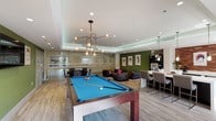 This amenity space is thoughtfully designed to offer both indoor recreation and outdoor relaxation. The interior game room features a striking tray ceiling with ambient LED cove lighting and a modern multi-arm chandelier that illuminates a central billiards table with a vivid blue felt surface. Wood-look flooring runs throughout, complementing the contemporary aesthetic. A lounge area with bean bag seating and accent chairs sits against a bold green accent wall adorned with framed artwork, while a bar-height counter with seating provides a social gathering spot. Glass partitions separate an additional seating or gaming area, maintaining an open and connected feel.

The outdoor spaces are equally well-appointed. A paved patio features a built-in outdoor fireplace flanked by distinctive high-back woven lounge chairs, creating an intimate seating nook. Nearby, a generous sectional sofa surrounds a rectangular fire pit table, offering a communal gathering space. A dedicated ping-pong table sits amid tall palm trees with views of a resort-style pool beyond a wrought iron fence. Lush landscaping, mature palms, and manicured shrubs tie the outdoor areas together, creating an inviting and resort-like atmosphere throughout.
