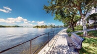 Located at 6116 Yellow Sun Drive in Lantana, Florida, this outdoor waterfront space offers a tranquil and picturesque setting along the edge of a calm, open lake. The space features a paved concrete promenade running parallel to the water's edge, bordered by a sturdy metal railing that provides both safety and an unobstructed sightline across the glistening lake. Mature, broad-canopy trees line the walkway, casting dappled shade over the path and contributing to a cool, natural atmosphere even under the bright South Florida sun. Wooden slatted benches are positioned along the promenade, offering comfortable spots to sit and take in the expansive water views. Across the lake, residential properties with docks and lush tropical landscaping are visible, enhancing the sense of a peaceful, established waterfront community. The well-maintained green lawn area between the walkway and the adjacent building adds a soft, park-like quality to the space. Under clear blue skies with scattered clouds, the area is bathed in abundant natural light, reflecting off the water's surface. This outdoor area serves as a calm retreat and community amenity, ideal for leisurely strolls, relaxation, and enjoying the natural beauty of Lantana's lakefront environment.