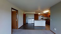 This single-floor space is designed with an open-concept layout that seamlessly connects the living area, kitchen, and utility zone. The kitchen is outfitted with rich, warm-toned wood cabinetry that extends to the ceiling, providing generous storage. A central island with a light-colored countertop offers additional workspace and serves as a natural focal point. Stainless steel appliances, including a microwave, electric range, and refrigerator, lend a modern touch to the kitchen. A pendant light hangs above the island, adding a subtle decorative element. Adjacent to the kitchen, a dedicated utility alcove houses a full-size washer and dryer alongside a water heater, all neatly enclosed within matching wood paneling for a cohesive look. The flooring transitions from dark hardwood in the kitchen and utility area to neutral carpeting in the living space. The overall color palette is understated, with off-white walls and warm wood tones creating a practical and comfortable environment. The layout maximizes functionality while maintaining an open, connected feel throughout the space.