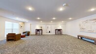 Located at 227 North Walnut in East Orange, NJ, this well-appointed single-floor multipurpose lounge offers a generously sized, open layout ideal for gatherings and flexible use. The space is finished with a distinctive patterned carpet featuring an abstract swirl design in neutral tones, which anchors the room and adds visual interest throughout.

The interior is divided into clearly defined functional zones. A comfortable seating area is centered around a bold red circular coffee table, surrounded by plush brown armchairs and accented by a tiered drum pendant light overhead. Natural light filters into this area through frosted windows, creating a warm, inviting ambiance. Framed artwork adorns the white walls, contributing to the space's refined aesthetic.

Along one wall, two high-top wooden desks with bar-height chairs are neatly arranged beneath wall-mounted lamps, offering dedicated workstation or consultation areas. A padded bench and additional wall art provide further finishing touches. The entry features sleek black-framed glass doors, and the space is equipped with a wall-mounted climate control unit and recessed ceiling lighting throughout. The clean white walls and smooth ceilings enhance the bright, open feel of the entire room, making it a versatile and professionally designed communal space.
