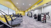 Located at 520 W Main St in downtown Oklahoma City, this fitness center offers a generously sized, single-floor workout facility designed to support a wide range of exercise routines. The space is defined by its bold yellow upper walls contrasted against light gray lower walls, creating an energetic and motivating atmosphere throughout. Recessed ceiling lighting provides bright, even illumination across the entire floor, which is finished with durable, patterned carpet tile in a neutral gray tone.

The layout is open and well-organized, accommodating a comprehensive selection of equipment. A dedicated cardio zone along one side features a row of treadmills and elliptical machines, while a strength training area is equipped with multiple cable and resistance machines, including multi-function cable stations and seated weight machines with padded seating. A free weights section includes a full dumbbell rack, kettlebells, and benches, accompanied by floor-to-ceiling mirrors that enhance the sense of space and allow users to monitor their form.

Additional functional training equipment includes rowing machines, a stair climber, medicine balls, resistance accessories, and storage shelving. A large mirrored wall runs along one side, further amplifying the open feel of the space. The facility also includes a utility storage closet and is served by a modern HVAC system with multiple ceiling vents ensuring comfortable air circulation throughout workouts.
