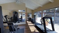 This fitness center offers a bright and welcoming environment designed to support a well-rounded workout routine. The space features soaring vaulted ceilings with exposed white-painted beams, lending an open and airy feel throughout. Large sliding glass doors and generous windows line the walls, flooding the room with natural light and providing views of the surrounding outdoor landscape.

The flooring consists of warm hardwood planks that extend across the main exercise area, complemented by rubber mat sections beneath the strength equipment for added safety and comfort. The equipment layout is thoughtfully arranged, with cardio machines — including treadmills and elliptical trainers — positioned along the windowed perimeter to allow users to enjoy outdoor views during their workouts. A multi-station cable and weight machine anchors the strength training area, offering a variety of resistance exercises.

Full-length mirrors are mounted along one wall, enhancing the sense of space while providing useful visual feedback during workouts. A ceiling fan helps maintain comfortable airflow. The overall ambiance is clean, functional, and well-lit, making this fitness center a practical and inviting space for daily exercise.