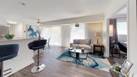 Located at 655 Americana Drive in Annapolis, Maryland, this single-floor residence offers a thoughtfully designed layout with contemporary finishes throughout. The open living and dining area is a standout feature, showcasing wide-plank hardwood-style flooring, sheer floor-to-ceiling vertical drape panels, and expansive sliding glass doors that flood the space with natural light and provide views of the surrounding greenery. A ceiling fan adds both function and style to the dining nook, which comfortably accommodates a glass-top table and chairs. The kitchen is efficiently designed with crisp white cabinetry, granite-style countertops, a decorative tile backsplash, and a full suite of stainless steel appliances including a gas range, built-in microwave, dishwasher, and refrigerator. The bedroom is a serene retreat featuring hardwood-style flooring, a large window dressed with patterned curtains and blinds, and ample space for a full dresser. A versatile secondary room serves as a functional home office, complete with a built-in shelving unit, closet, and a window with natural light. The bathroom is finished with a granite-topped vanity, white cabinetry, and an octagonal mirror, delivering a clean and polished aesthetic.