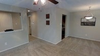 This single-floor residence offers a straightforward and functional layout designed for comfortable everyday living. The main living area is finished with wall-to-wall carpet in a neutral tone, creating a warm and inviting atmosphere. A ceiling fan with a light fixture is centrally positioned overhead, providing both ventilation and ambient lighting. The textured ceiling adds a classic touch to the space. Neutral gray walls keep the interior feeling clean and versatile, accented by two decorative wall-mounted panels that add a subtle artistic element. The living area flows openly toward the kitchen, which is visible through a wide doorway and features dark wood cabinetry, offering a contrast to the lighter tones of the main living space. A hallway extends from the living area, lined with recessed lighting and leading to additional rooms and a exterior door, suggesting a well-connected floor plan. White baseboards and trim throughout the home provide a crisp finish that ties the design together cohesively. Overall, the space presents a practical and well-maintained interior with a neutral palette suited to a variety of furnishing styles.