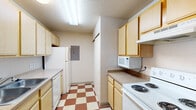 Located on Collegiate Way in Mount Pleasant, Michigan, this single-floor unit offers three bedrooms and two full bathrooms within a efficiently organized floor plan. The space is designed to maximize functionality across all rooms.

The galley-style kitchen features light wood cabinetry on both sides, a dual-basin sink, a white electric range, dishwasher, refrigerator, and a built-in microwave. The distinctive red-and-cream checkerboard vinyl flooring adds a notable visual element, while a textured ceiling and recessed lighting complete the space.

The living area provides access to an exterior door and windows that allow natural light to filter in. Carpeted flooring runs throughout the living, dining, and bedroom areas. The dining space accommodates a round table and chairs. The bedrooms include closets with sliding mirrored doors, windows for natural light, and built-in desk shelving units in some rooms.

The bathrooms include a full tub-shower combination, tile flooring, and wood-toned vanity cabinetry. Overall, the unit presents a straightforward, functional layout suited for everyday living.