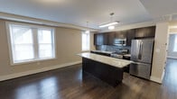 This single-floor residence offers a well-designed open-concept layout that seamlessly blends the kitchen and living areas into one cohesive, functional space. The kitchen is a standout feature, showcasing rich dark cabinetry that extends to the upper walls, providing generous storage. A large central island with light granite countertops serves as the heart of the kitchen, ideal for meal preparation or casual dining. The countertops continue throughout the kitchen, complementing the elegant subway tile backsplash in a soft gray tone. Stainless steel appliances — including a gas range, microwave, and refrigerator — complete the modern culinary setup. Pendant lighting hangs above the island and along the kitchen perimeter, offering warm, focused illumination. The hardwood flooring, finished in a deep espresso tone, flows throughout the open living space, adding warmth and visual continuity. Multiple windows allow natural light to fill the room, enhancing the airy, open atmosphere. Neutral-toned walls provide a clean backdrop that complements the bold cabinetry and flooring, creating a balanced, contemporary aesthetic throughout the space.