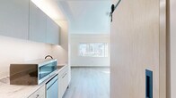 Located on the eighth floor at 361 Turk Street in San Francisco's Tenderloin neighborhood, this efficiently designed studio unit offers a thoughtfully organized single-level layout. The open living area features light wood-tone flooring throughout, crisp white walls, and a wall-mounted flat-screen television, creating a clean and functional living environment. A wide window frames urban city views, filling the space with natural light and providing an engaging connection to the San Francisco skyline.

The integrated kitchenette is positioned along one wall and includes light-toned cabinetry, a compact countertop with sink, microwave, and under-counter appliances — all designed to maximize utility within a minimal footprint. A sliding barn-style door with a frosted glass panel separates zones within the unit, adding both practicality and a modern aesthetic touch.

The bathroom is designed with accessibility in mind, featuring large-format light tile flooring, a fully open roll-in shower with matte black grab bars and a handheld showerhead, a wall-mounted sink with an illuminated mirror cabinet above, and a wall-hung toilet. A light wood storage bench within the shower area adds a functional and warm detail. The overall design of the unit prioritizes clean lines, accessibility, and modern minimalism throughout.