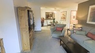 This single-floor residence offers an open and cohesive layout that seamlessly connects the living and dining areas, creating a welcoming and functional flow throughout the home. The living area is anchored by plush wall-to-wall carpeting in a warm neutral tone, complemented by a substantial rustic wooden entertainment armoire that adds character and warmth to the space. Comfortable upholstered seating, including a patterned accent chair with a decorative pillow, invites relaxation around a sleek dark coffee table. Moving through the space, the dining area is outfitted with a round table and elegant wrought-iron style chairs, set beneath a ceiling fan with light fixture. A decorative mirror with an ornate frame is mounted on the wall, enhancing the sense of depth and light in the room. Tasteful framed artwork and potted floral arrangements add color and personality throughout. The neutral wall palette keeps the overall ambiance calm and airy, while the consistent carpeting unifies the open floor plan. Additional rooms are accessible from the main living space, suggesting a practical and well-organized layout designed for comfortable everyday living.