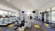 This single-floor fitness center is designed to support a full range of workout routines in a bright, open environment. The main workout area features soaring ceilings with recessed lighting and is flooded with natural light through large windows and an elegant arched window, offering pleasant outdoor views of the surrounding greenery and community. The flooring throughout consists of durable commercial carpet tiles in a distinctive pattern of gray, yellow, and blue, lending the space a modern, energetic aesthetic.

The facility is comprehensively equipped with multiple strength training cable machines, weight benches, a squat rack with free weights, and a full dumbbell rack with stability balls and resistance accessories. A dedicated cardio zone lines the perimeter, featuring a row of treadmills positioned directly in front of floor-to-ceiling windows for an inspiring workout experience. Additional cardio options include stationary bikes and a rowing machine. A separate functional training area provides TRX suspension straps and a pull-up rig for bodyweight and resistance-band workouts, along with spin bikes.

The entry area transitions from tile flooring and includes a water fountain station, a fire safety cabinet, and restroom access. Mounted flat-screen TVs are positioned throughout the main floor for entertainment. The space is clean, well-maintained, and thoughtfully laid out to accommodate a variety of fitness goals.