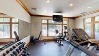 Located at 1718 E Lincoln Rd in Spokane, WA, this fitness center offers a well-equipped exercise space designed to support a range of workout routines. The room features durable rubber flooring throughout, providing a safe and stable surface for both cardio and strength training activities. A textured ceiling with recessed lighting and warm wood crown molding gives the space a polished, finished look.

The facility is outfitted with a comprehensive selection of cardio equipment, including treadmills, a StairMaster, an elliptical, and a stationary bike. A dedicated free weights area features a full rack of dumbbells, while a wall-mounted resistance station provides additional strength training options. A large wall mirror runs along one side of the room, enhancing the sense of space and allowing users to monitor their form during workouts.

Natural light filters in through multiple wood-framed windows, offering views of the surrounding outdoor area and creating a bright, energizing atmosphere. A wall-mounted flat-screen television adds entertainment value for users during cardio sessions. Posted fitness rules ensure the space remains organized and welcoming for all users. A wood-framed glass entry door with a transom window completes the space, contributing to the overall warm and functional design aesthetic.