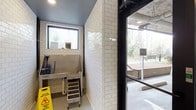 This single-floor space includes a purpose-built pet grooming area designed with both functionality and cleanliness in mind. The washing station is lined with classic white subway tiles that extend from floor to ceiling, creating a bright, hygienic environment that is easy to maintain. A deep stainless steel wash tub serves as the centerpiece, equipped with a mounted hose and a small access step stool, making it convenient for bathing pets of various sizes. A window above the tub allows natural light to filter in, offering views of the surrounding wooded landscape. The ceiling is painted a soft gray-blue, adding a subtle design touch to the utilitarian space. A caution wet floor sign indicates the area is actively used and well-maintained. A dark-framed glass door opens directly to an outdoor area, where a landscaped courtyard with steps, seating, and mature trees is visible, providing a seamless transition between the interior washing station and the exterior environment. The overall design prioritizes practicality while maintaining a clean, polished aesthetic suited to a shared amenity space.