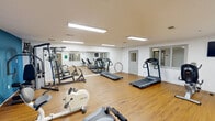 This fitness center in Alexandria, VA offers a well-equipped, single-floor exercise facility designed to support a range of workout routines. The space features warm hardwood flooring throughout, crisp white walls accented by a bold blue feature wall, and bright fluorescent lighting that ensures excellent visibility across the entire floor.

The cardio area is furnished with multiple treadmills and stationary bikes, providing ample options for aerobic exercise. A large wall mirror spans one side of the main workout area, enhancing the sense of openness and allowing users to monitor their form. A wall-mounted flat-screen TV is positioned for easy viewing during workouts.

The strength training section includes a multi-function cable and weight machine, a freestanding weight rack, and an adjustable workout bench, catering to a variety of resistance training needs. Additional equipment such as exercise hoops rounds out the functional fitness offerings.

The facility includes an interior window pass-through and a separate door that connects to an adjacent room, suggesting additional support spaces. Ventilation is provided via ceiling-mounted HVAC vents, and safety is addressed with a visible fire alarm on the wall. The overall layout is open and functional, providing a practical and comfortable environment for daily fitness activities.