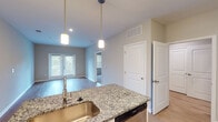 This single-floor residence is designed with a clean, modern aesthetic that balances functionality and style throughout. The open-concept kitchen and living area form the heart of the unit, where light hardwood-style flooring extends seamlessly across the space. The kitchen is well-appointed with white shaker-style cabinetry reaching toward the ceiling, granite countertops, a stainless steel appliance suite including a refrigerator, range, microwave, and dishwasher, and a tiled backsplash in a soft blue-green hue. A granite-topped island with a stainless steel sink and pendant lighting above serves as a natural divider between the kitchen and the open living area. French doors in the living space allow natural light to filter in and provide direct access to an outdoor balcony overlooking greenery. A separate bedroom features plush carpeting, a ceiling fan, and a standard window for natural light. An additional room with hardwood-style flooring and a sliding glass door opens directly to the balcony. The bathroom includes a granite vanity with white cabinetry and a lighted mirror, with an adjacent walk-in closet fitted with wire shelving. Neutral warm-toned walls and consistent finishes create a cohesive ambiance throughout.