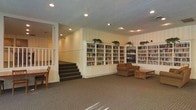 This communal library and lounge space offers a warm, functional environment designed for relaxation and reading. The room is anchored by an expansive wall of white built-in bookshelves filled with a wide variety of books, extending nearly the full length of the space. Potted plants are positioned atop the shelving units, adding touches of greenery and life to the area. Recessed lighting in the textured ceiling provides even, comfortable illumination throughout.

A cozy seating arrangement, including an upholstered armchair and a Mission-style wood coffee table, is situated near the bookshelves, creating an inviting reading nook. A television is integrated into the shelving wall, adding a media component to the lounge. The floor is covered in neutral-toned carpet, contributing to the room's quiet, comfortable atmosphere.

A carpeted staircase with a white railing is visible to one side, indicating access to another level. Additional seating with a simple wooden chair and table can be found nearby. The walls feature subtle striped wallcovering in soft tones, enhancing the space's understated, classic aesthetic. Overall, the space feels organized, serene, and purposefully designed for communal use.