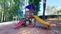 This outdoor playground offers a welcoming recreational space set within a natural, tree-lined environment. Towering pines and mature deciduous trees provide generous shade across the grounds, creating a cool and comfortable setting for active play.

At the heart of the space stands a multi-level wooden play structure, constructed with sturdy timber framing finished in bold, contrasting colors. The central tower features a purple canopy overhead, offering additional shade on the upper platform. Two slides extend from the structure — a red slide on the front face and a bright yellow slide on the side — providing multiple options for descent and encouraging active movement.

Adjacent to the main structure, a swing set is visible toward the rear of the play area, expanding the range of activities available. The ground throughout the space is covered in a natural wood chip surface, which provides cushioning underfoot and blends seamlessly with the surrounding woodland setting.

The overall atmosphere is lively yet naturally serene, with dappled sunlight filtering through the tree canopy above. The combination of colorful equipment and a verdant, wooded backdrop makes this playground an engaging and visually appealing outdoor destination well-suited for energetic, imaginative play.