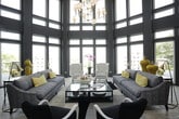 This single-story space presents a cohesive and sophisticated interior defined by a striking contrast of dark navy cabinetry, crisp white surfaces, and warm gold hardware and pendant lighting. The design aesthetic is consistent throughout, blending modern functionality with bold decorative choices.

The kitchen area is fully equipped with stainless steel appliances, including a range with a custom hood, built-in microwave, and a coffee station. Dark shaker-style cabinetry reaches toward the ceiling, complemented by white subway tile backsplashes and light wood-look flooring that runs throughout the space.

A wet bar or secondary kitchenette features an undermount sink, open shelving, and gold pendant lights overhead, offering additional utility and style. Adjacent to these areas, a lounge space provides a comfortable seating area anchored by a linear fireplace with white brick surround and a wall-mounted television. Oversized windows flood the lounge with natural light and offer exterior views.

A separate sitting nook showcases a bold patterned sectional sofa and abstract wall art, adding an artistic, eclectic touch. The leasing or reception area features a dramatic dark desk, glass-partitioned conference space, and a mounted display screen, rounding out this well-appointed, multi-functional space.
