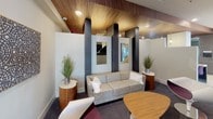 This thoughtfully designed single-floor amenity space blends contemporary style with functional versatility. The entrance foyer greets visitors with large floor-to-ceiling windows that flood the space with natural light, light-toned tile flooring, and a striking chevron-patterned accent wall accented by a bold yellow logo sign. The open layout flows seamlessly between distinct zones, each with its own character and purpose.

The lounge area features plush gray sofas, a rounded wood coffee table, and modern side tables flanked by decorative greenery. Bold dark columns rise to a warm wood-paneled ceiling, adding architectural depth, while curated wall art lends a polished, inviting ambiance. Carpeted flooring and recessed lighting complete the comfortable setting.

A dedicated conference room offers a sleek oval wood table surrounded by ergonomic chairs, a wall-mounted flat-screen display, and elegant pendant and sconce lighting, with a window providing garden views — ideal for professional meetings.

The wet bar area is a design highlight, featuring wood cabinetry, a white countertop, stainless steel appliances, and a dazzling metallic penny-tile backsplash with integrated open shelving. Nearby, a game lounge with a pool table provides a social gathering space. A tufted gold banquette seating nook with patterned walls adds a luxurious, lounge-like feel. Wide corridors with wood-paneled ceilings and framed artwork connect all areas cohesively.