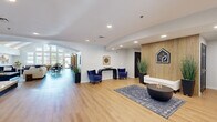 This thoughtfully designed single-floor amenity space offers a generous and well-appointed environment for residents to gather, work, and stay active. The welcoming lobby entrance features light hardwood flooring, clean white walls, vertical wood slat paneling, and a branded feature wall, setting a polished and contemporary tone. Plush navy accent chairs, a console table, and curated greenery create an inviting first impression.

The expansive lounge area showcases soaring vaulted ceilings with illuminated beam details, a striking floor-to-ceiling fireplace, a large wall-mounted display, and ample seating for relaxation and socializing. Tall windows flood the space with natural light and offer views of the surrounding outdoor landscape.

A dedicated dining and social zone features a combination of booth-style seating, communal tables with pendant lighting, and a sleek built-in refreshment bar with dark navy cabinetry, gold hardware, a patterned green tile backsplash, and open shelving — perfect for casual gatherings.

A private meeting room, carpeted and adorned with abstract wall art, provides an intimate setting for small group discussions. The fully equipped fitness center includes treadmills, an elliptical, stationary bikes, and interactive mirror fitness systems, all beneath a geometric LED light fixture, with natural light from surrounding windows completing the energizing atmosphere.