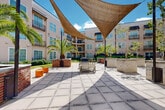 Located at 4301 Tulane Ave in New Orleans, Louisiana, this outdoor courtyard space serves as a central gathering area within a well-appointed multi-story residential complex. The courtyard is defined by its broad, grid-patterned concrete pavers that create a clean and open plaza ideal for relaxed outdoor living. Overhead, large triangular shade sails are stretched between poles, providing ample shelter from the New Orleans sun while adding a contemporary design element to the space. Lush tropical landscaping is woven throughout, with mature palm trees planted in raised brick-clad planters serving as striking focal points. Neatly trimmed hedges, flowering shrubs, and planted garden beds line the perimeter, softening the transition between the hardscape and the surrounding building facade. Seating areas with benches and casual furniture are positioned throughout the courtyard, encouraging social interaction and relaxation. The surrounding multi-story building features neutral-toned siding, large windows, and private balconies with wrought-iron railings that overlook the courtyard below. Pedestrian walkways weave through manicured green lawns along the building's edges, connecting various entries and adding to the overall accessibility and flow of the outdoor environment. The space enjoys abundant natural light and an open-sky ambiance, reflecting the warm, vibrant character of New Orleans.