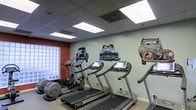 This fitness center is designed to keep guests and residents active while offering a visually engaging environment. The space is outfitted with a selection of modern cardio equipment, including multiple treadmills with individual display screens and an elliptical trainer, providing a comprehensive workout experience. A standout design element is the eye-catching wall mural depicting vintage classic cars in varying states of wear, adding a retro, artistic character to the room. The walls feature a complementary color palette of soft sage green and bold terracotta accents, contributing to a lively yet comfortable atmosphere. Natural light filters in through a glass block window on one side, supplemented by bright recessed panel lighting in the drop ceiling above, ensuring the space remains well-lit at all times. The flooring and layout are practical and functional, allowing for ease of movement between equipment. Overall, this fitness center balances utility with creative interior design, making it an inviting space for a workout.