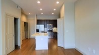 This single-floor space centers around an open-concept kitchen that serves as the heart of the layout. The kitchen is equipped with a full suite of stainless steel appliances, including a French door refrigerator, a microwave, and a gas range, all framed by rich dark cabinetry that extends to the upper walls. A decorative tile backsplash adds visual interest behind the range, complementing the overall color palette. A white kitchen island with a light-toned countertop provides additional workspace and storage, functioning as a natural dividing point between the kitchen and the adjacent living areas. Warm hardwood flooring flows continuously throughout the space, adding depth and cohesion. Recessed lighting is distributed evenly across the ceiling, ensuring consistent, functional illumination throughout. The clean white walls and neutral tones create a bright, airy atmosphere. Multiple doorways visible from the main area suggest well-defined transitions to additional rooms, contributing to a practical and organized floor plan overall.