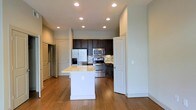 This single-floor space centers around an open-concept kitchen that serves as the heart of the layout. The kitchen is equipped with a full suite of stainless steel appliances, including a French door refrigerator, a microwave, and a gas range, all framed by rich dark cabinetry that extends to the upper walls. A decorative tile backsplash adds visual interest behind the range, complementing the overall color palette. A white kitchen island with a light-toned countertop provides additional workspace and storage, functioning as a natural dividing point between the kitchen and the adjacent living areas. Warm hardwood flooring flows continuously throughout the space, adding depth and cohesion. Recessed lighting is distributed evenly across the ceiling, ensuring consistent, functional illumination throughout. The clean white walls and neutral tones create a bright, airy atmosphere. Multiple doorways visible from the main area suggest well-defined transitions to additional rooms, contributing to a practical and organized floor plan overall.
