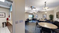 This single-floor community space is thoughtfully designed to accommodate a variety of social and professional gatherings. The main area functions as a generous dining and lounge room, furnished with multiple round tables and padded chairs arranged to foster comfortable group interaction. Overhead, a combination of ceiling fans and pendant light fixtures provide both functional illumination and a welcoming ambiance, while patterned carpet tiles add texture underfoot. Framed artwork and a decorative indoor plant contribute to the warm, hospitable atmosphere. Windows fitted with horizontal blinds allow for adjustable natural light throughout the space.

A separate, more intimate conference or meeting room branches off from the main area, offering a quieter setting for smaller gatherings or private discussions. This room features its own round table with seating for several, a wall-mounted flat-screen television, a cork bulletin board, and pendant lighting. A window with blinds provides natural light, and decorative framed prints add a polished touch.

Serving both areas is a well-appointed kitchen, visible through a pass-through counter with a laminate surface. The kitchen is equipped with rich wood cabinetry, stainless steel appliances including a refrigerator, range, microwave, and dishwasher, and a tasteful tile backsplash, making it ideal for catering or communal meal preparation.