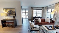 This single-floor residence showcases a thoughtfully designed open-concept layout that seamlessly connects the living, dining, and kitchen areas. The living space is anchored by warm hardwood-style flooring that extends throughout, complementing the neutral gray walls and crisp white trim for a clean, contemporary aesthetic.

Large windows dressed with patterned drapery panels flood the interior with generous natural light while offering views of the surrounding urban streetscape. The living area comfortably accommodates a rich cognac leather sofa paired with modern upholstered accent chairs arranged around a patterned area rug in deep blues and earth tones, creating a welcoming focal point.

A defined corridor connects the living space to what appears to be a kitchen and additional areas beyond, where dark cabinetry provides a striking contrast against the lighter surroundings. The dining area sits adjacent to the main windows, taking advantage of the natural light.

Overall, the space presents a cohesive blend of modern and transitional design elements, combining warm textures, curated decor, and an open flow that makes the interior feel both functional and inviting.