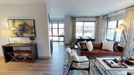 This single-floor residence showcases a thoughtfully designed open-concept layout that seamlessly connects the living, dining, and kitchen areas. The living space is anchored by warm hardwood-style flooring that extends throughout, complementing the neutral gray walls and crisp white trim for a clean, contemporary aesthetic.

Large windows dressed with patterned drapery panels flood the interior with generous natural light while offering views of the surrounding urban streetscape. The living area comfortably accommodates a rich cognac leather sofa paired with modern upholstered accent chairs arranged around a patterned area rug in deep blues and earth tones, creating a welcoming focal point.

A defined corridor connects the living space to what appears to be a kitchen and additional areas beyond, where dark cabinetry provides a striking contrast against the lighter surroundings. The dining area sits adjacent to the main windows, taking advantage of the natural light.

Overall, the space presents a cohesive blend of modern and transitional design elements, combining warm textures, curated decor, and an open flow that makes the interior feel both functional and inviting.