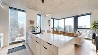 Located at 160 N Morgan Street in Chicago's vibrant West Loop neighborhood, this contemporary high-rise unit delivers a compelling blend of industrial and modern design. Exposed concrete ceilings throughout lend an urban loft aesthetic, while warm wood flooring and crisp white walls create a bright, refined atmosphere.

The open-concept kitchen and living area form the heart of the space. The kitchen is outfitted with light wood and navy cabinetry, stainless steel appliances, a large waterfall-edge island with bar seating, and a matte black pendant light fixture. Floor-to-ceiling windows wrap around the corner, flooding the space with natural light and offering panoramic views of the Chicago skyline.

The living room continues the window-forward design, framing sweeping city vistas and providing an airy, light-filled setting. The bedroom features a corner glass window configuration delivering unobstructed elevated views of the surrounding cityscape, complemented by the same exposed concrete ceiling detail. The bathroom is finished with a floating wood vanity, an illuminated mirror, a glass-enclosed walk-in shower, and neutral tile flooring. Throughout the unit, thoughtful design details and high-end finishes contribute to a polished, contemporary urban living experience.