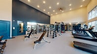 This single-floor fitness center is a thoughtfully designed and comprehensively equipped facility that caters to a wide range of workout needs. The space is divided into distinct functional zones, each offering a unique training environment. The main strength training area commands attention with soaring ceilings, dramatic dark accent walls, and recessed lighting that illuminates an extensive collection of free weights, adjustable benches, cable machines, and multi-use weight racks. Full-length mirrors line the walls, enhancing the sense of openness throughout. A dedicated cardio zone features treadmills and stationary bikes positioned in front of large floor-to-ceiling windows, flooding the area with natural light and providing outdoor views during workouts. Wall-mounted flat-screen TVs are conveniently placed for entertainment. A separate stretching and functional training area offers open floor space alongside stability balls, step platforms, and resistance equipment, while built-in teal shelving provides organized storage. A striking botanical-patterned wallcovering in deep teal tones adds a bold design statement near the entry, complementing the facility's modern, upscale aesthetic. The overall ambiance is energetic yet polished, making this fitness center a standout amenity.