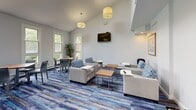 Located at 1037 Southern Artery in Quincy, Massachusetts, this single-floor community lounge space offers a thoughtfully designed environment ideal for socializing and relaxation. The open layout is unified by a distinctive blue, multi-toned striped flooring that lends the room a vibrant yet cohesive aesthetic.

The main lounge area features generous seating arrangements, including comfortable gray upholstered sofas and armchairs accented with bold blue patterned throw pillows. Wooden coffee tables and side tables are positioned throughout for convenience. A wall-mounted flat-screen television serves as a central focal point, making the space suitable for both casual gatherings and communal viewing.

A raised bar counter with white high-back stools occupies one side of the room, complemented by additional dining-height tables and chairs, providing flexible options for dining or working. Multiple large double-hung windows flood the space with natural light and offer pleasant views of the surrounding greenery.

The soaring ceiling height adds a sense of openness and grandeur, while pendant drum light fixtures provide warm, even illumination. Framed artwork is displayed along the crisp white walls, adding a polished, welcoming character. A hallway corridor connects to additional areas, decorated with coordinating artwork and recessed lighting, maintaining the clean, modern aesthetic throughout.