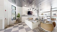 Located at 1153 Bees Ferry Rd in Johns Island, South Carolina, this single-floor community amenity space offers a generously proportioned layout designed for both relaxation and social gathering. The interior is defined by striking shiplap ceilings with a gray-painted finish, from which clusters of glass pendant lights and oversized ceiling fans are suspended, creating a coastal-modern aesthetic throughout.

The main lounge area centers around a sleek, linear gas fireplace set into a white shiplap accent wall, which also features a wall-mounted television above a gray slate mantle. Plush white sofas and woven rattan accent chairs are arranged around a vintage-style area rug, offering a warm and inviting seating arrangement. The signature checkerboard tile flooring — alternating soft gray and cream large-format tiles — extends throughout the entire space, lending a sophisticated yet relaxed character.

Large black-framed glass doors and transom windows line the perimeter, flooding the interior with abundant natural light and providing seamless transitions to the outdoor areas. A covered outdoor lounge features a hanging swing sofa, additional seating, and direct views of the resort-style pool beyond the black iron fencing. A bistro-style bar counter and dining area complete the space, making it a fully functional social hub for the surrounding community.