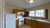 This single-floor space offers a straightforward and functional layout designed for comfortable everyday living. The living area features neutral wall tones, plush carpeting, and smooth ceilings, creating a clean, understated ambiance. A floor vent provides climate control, and a doorway with warm wood framing connects to an adjacent area, hinting at a practical flow between rooms.

The kitchen is well-equipped and thoughtfully arranged, featuring warm honey-toned wood cabinetry that extends from lower to upper sections, offering generous storage throughout. A white gas range sits beneath an overhead ventilation hood, complemented by a double-basin sink positioned along the countertop beneath a wide, multi-pane window that lets in ample natural light and provides views of the surrounding trees. A full-size white refrigerator anchors one end of the kitchen, while dark tile backsplash accents add visual contrast to the light countertops. The tile flooring in the kitchen provides a practical, easy-to-clean surface. Together, these spaces present a well-maintained, move-in ready environment with classic finishes that provide a solid foundation for personalization.